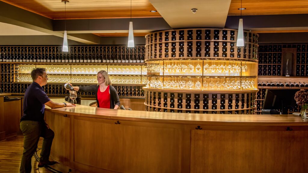 Reception area with a smiling staff member in a red shirt, surrounded by shelves filled with neatly arranged bottles. The warm wooden decor enhances the inviting atmosphere of the establishment.