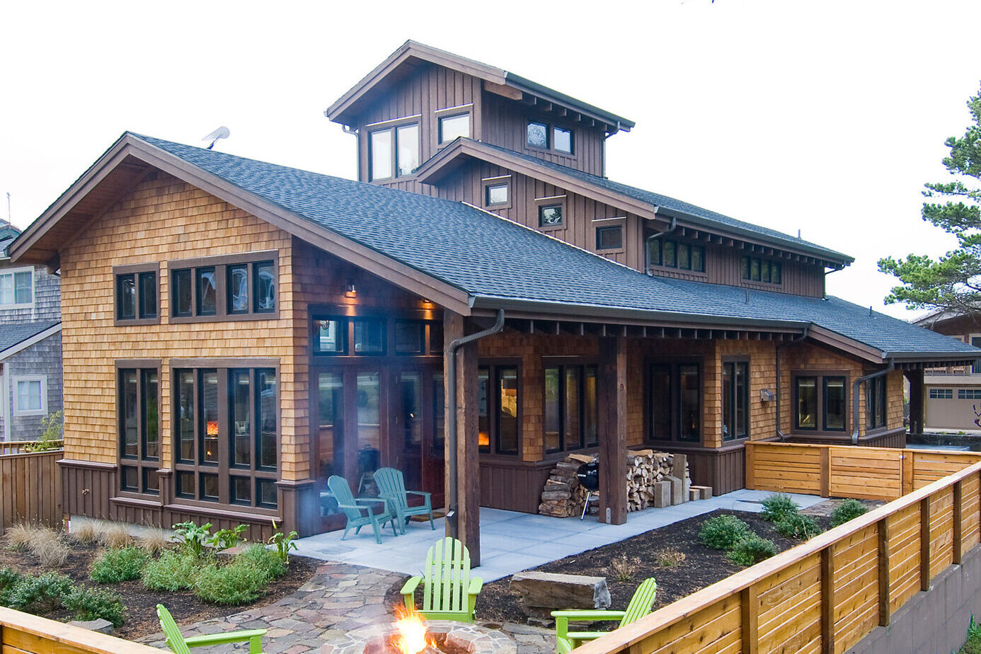 Modern wooden house with a spacious porch, large windows, and a cozy outdoor seating area, surrounded by landscaped greenery and a fire pit.