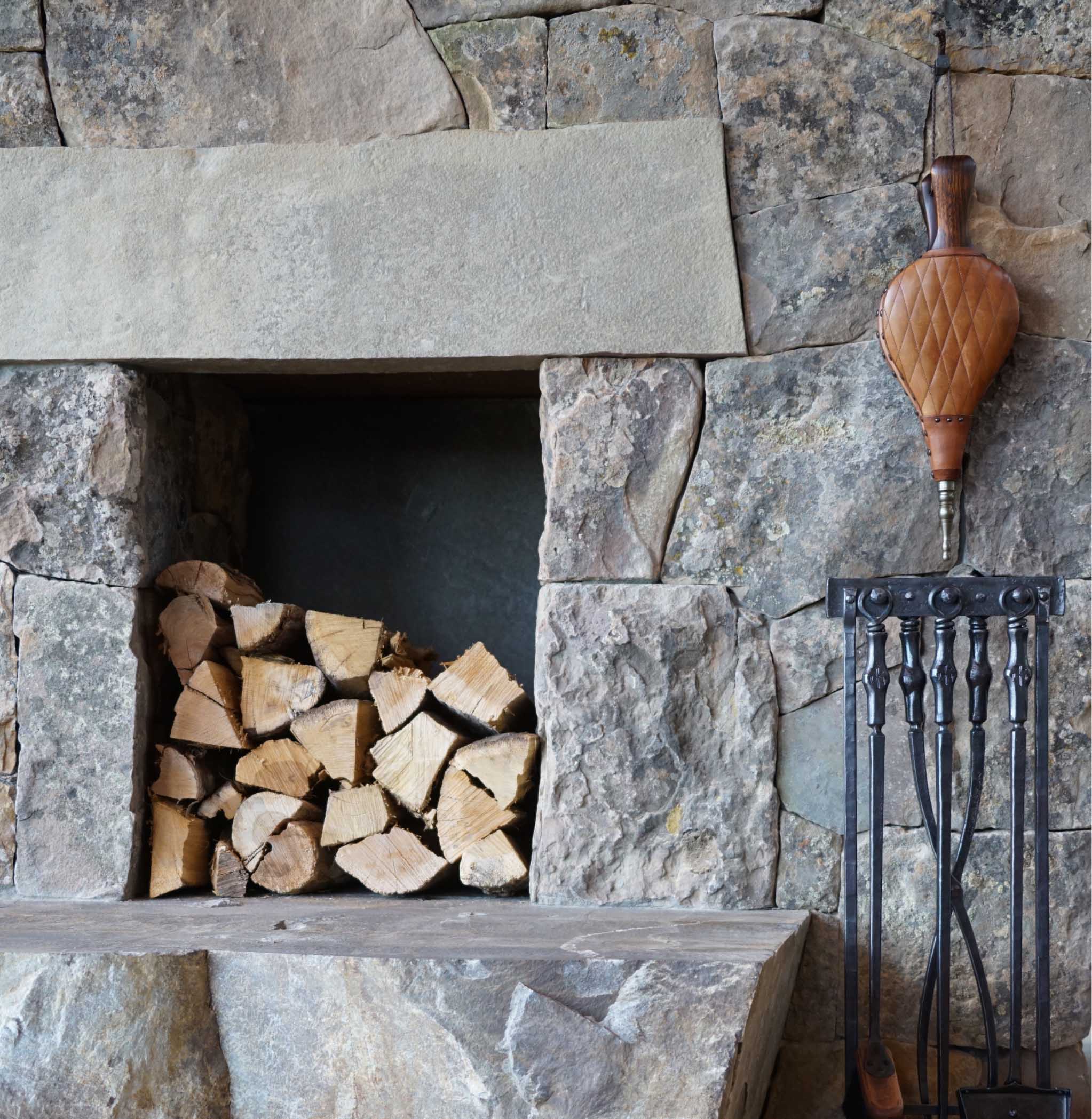 Rustic stone fireplace with neatly stacked firewood and a decorative wooden tool holder on the wall, creating a cozy, warm atmosphere.