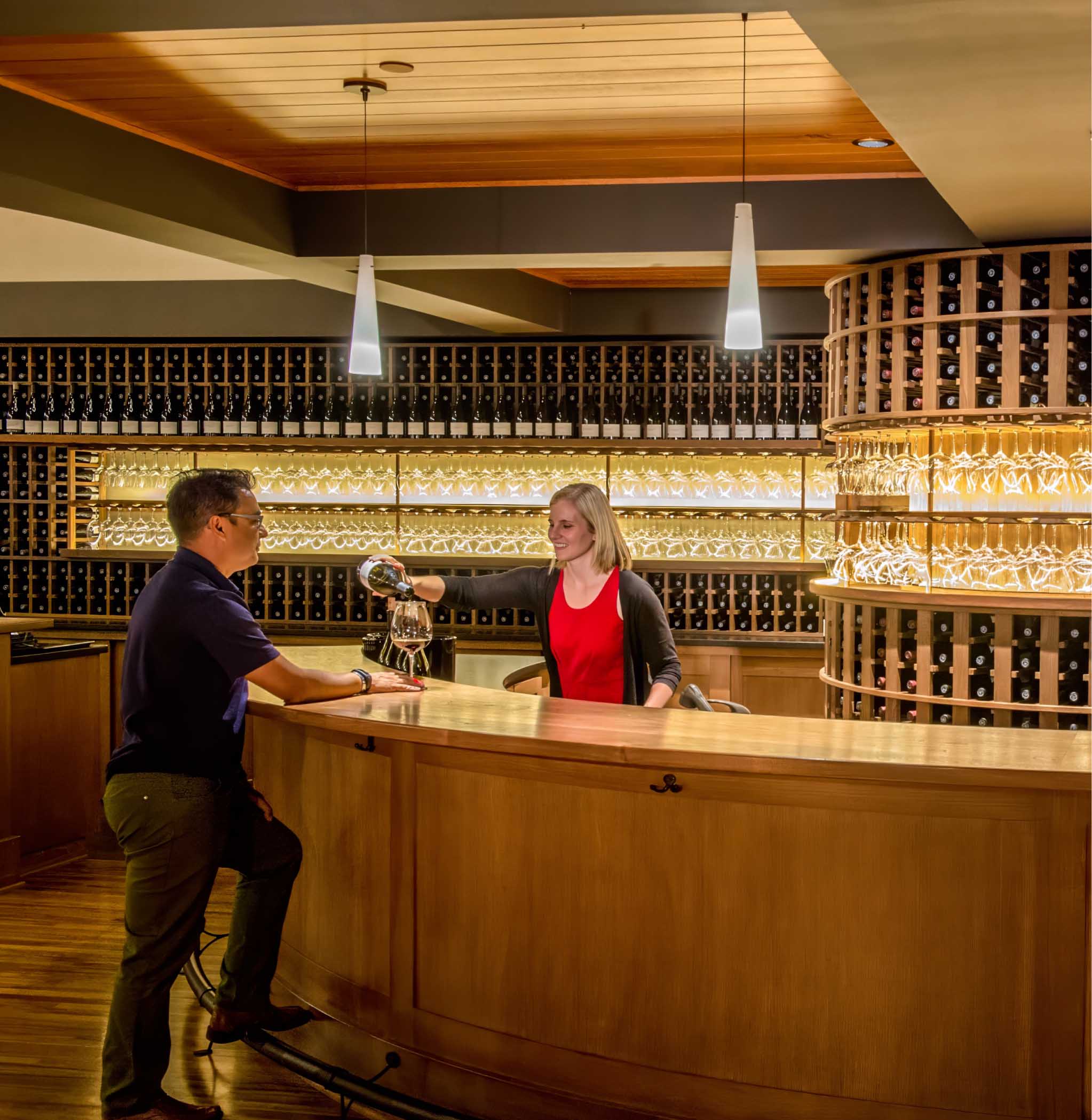 Reception area with a wooden counter, featuring a staff member in a red shirt assisting a guest. The background showcases a well-organized display of wine bottles, creating a warm and inviting atmosphere. Ideal for a wine tasting or hospitality setting.