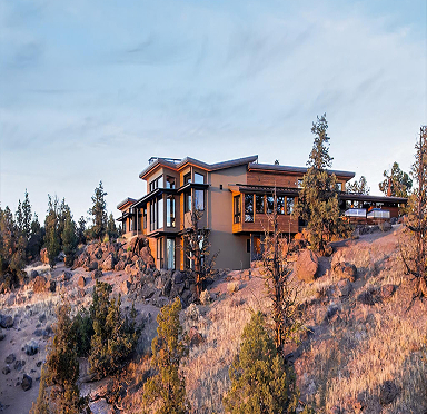 Modern hillside home with large windows and wooden deck, surrounded by trees and rocky terrain under a clear sky.
