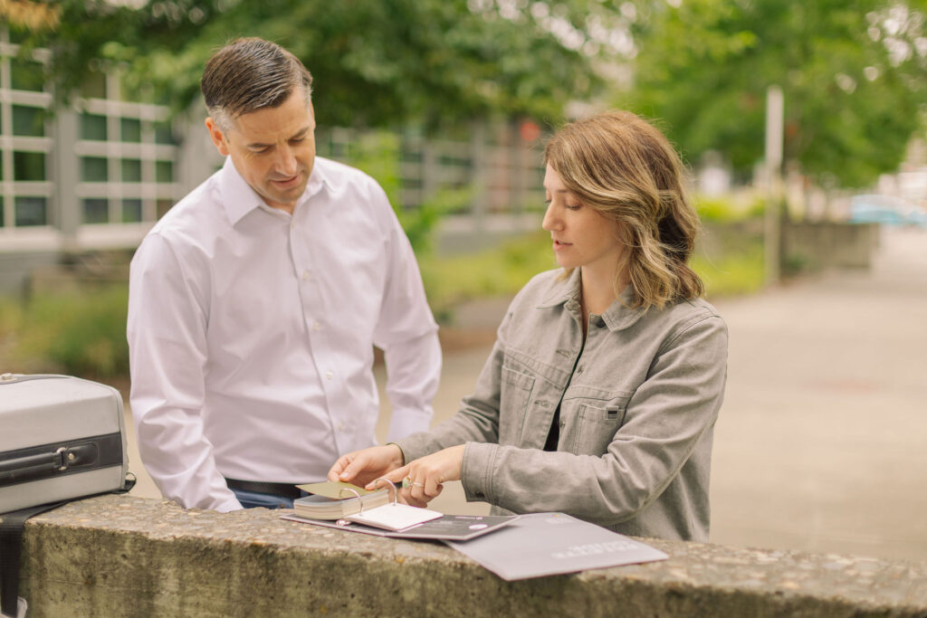 Business professionals discussing project details outdoors, with a focus on documents and planning materials. The scene features a man in a white shirt and a woman in a gray jacket, engaged in a collaborative conversation at a stone table.