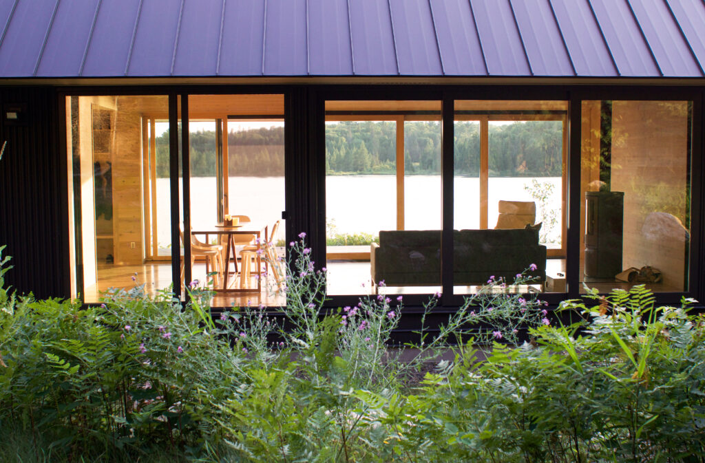 Modern lakeside cabin with large glass windows, showcasing a cozy interior featuring a dining table and sofa, surrounded by lush greenery and a serene lake view. Ideal for nature retreats and relaxing getaways.