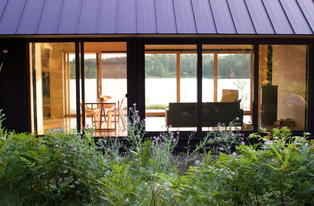 Modern lakeside cabin with large glass windows, showcasing a cozy interior featuring a dining table and sofa, surrounded by lush greenery and a serene lake view. Ideal for nature retreats and relaxing getaways.