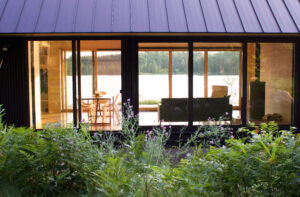 Modern lakeside cabin with large glass windows, showcasing a cozy interior featuring a dining table and sofa, surrounded by lush greenery and a serene lake view. Ideal for nature retreats and relaxing getaways.