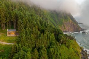 Aerial view of a modern cabin nestled among lush evergreen trees on a coastal cliff, with fog rolling over the mountains and the ocean visible in the background. The winding path leading to the cabin enhances the serene natural setting.