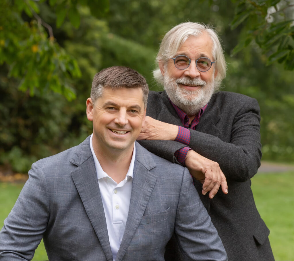 Nathan Good and Forrest Good pose together outdoors in a park setting, with one man seated in a light gray blazer and white shirt, smiling at the camera. The other man stands behind him, resting his hands on the seated man's shoulders, wearing a dark blazer and glasses, also smiling. The background features green foliage, suggesting a pleasant, natural environment.